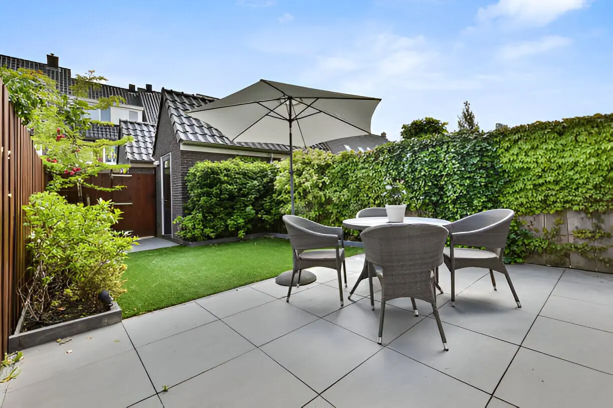Compact backyard patio with wicker dining set, patio umbrella, artificial turf lawn, and lush green hedge privacy screening