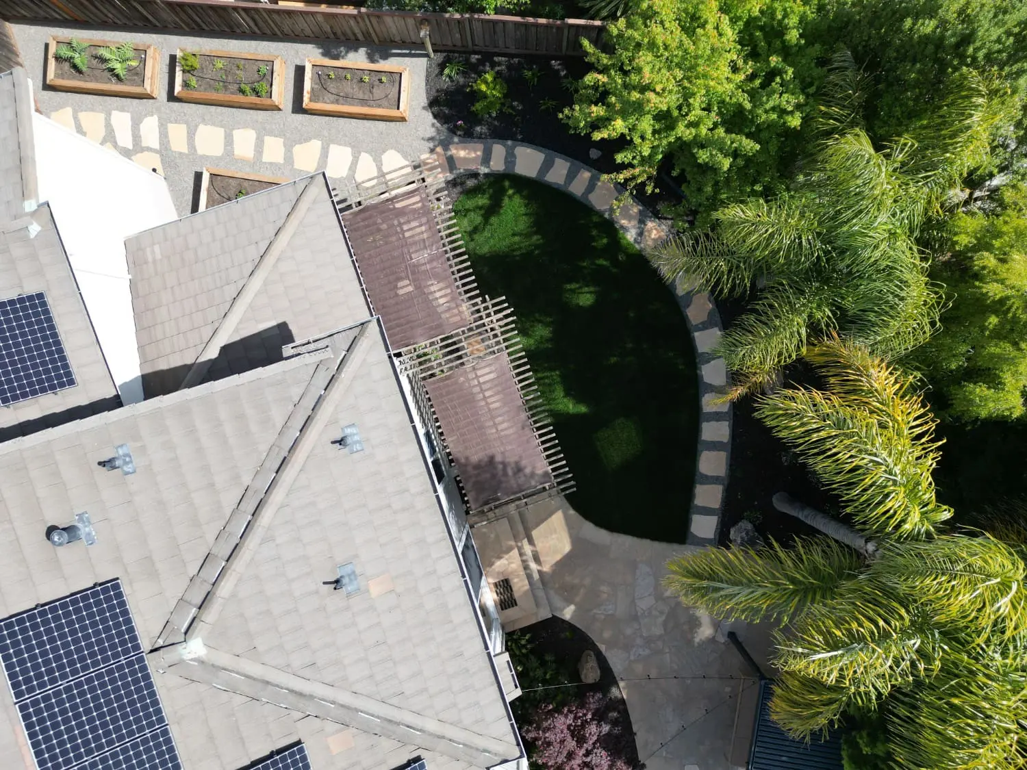 Aerial view of a solar-paneled home with a curved turf lawn bordered by a stone edge, a wood pergola, and raised garden beds on a gravel patio, an example of landscape design in Oakville, CA by Keystone Yards.