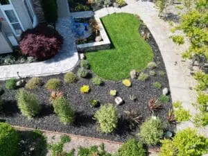 Aerial view of a landscaped side yard by Keystone Yards, with a curved lawn, mulched planting beds of ornamental grasses and succulents, a paver patio with blue Adirondack chairs, and a Japanese maple by the house.