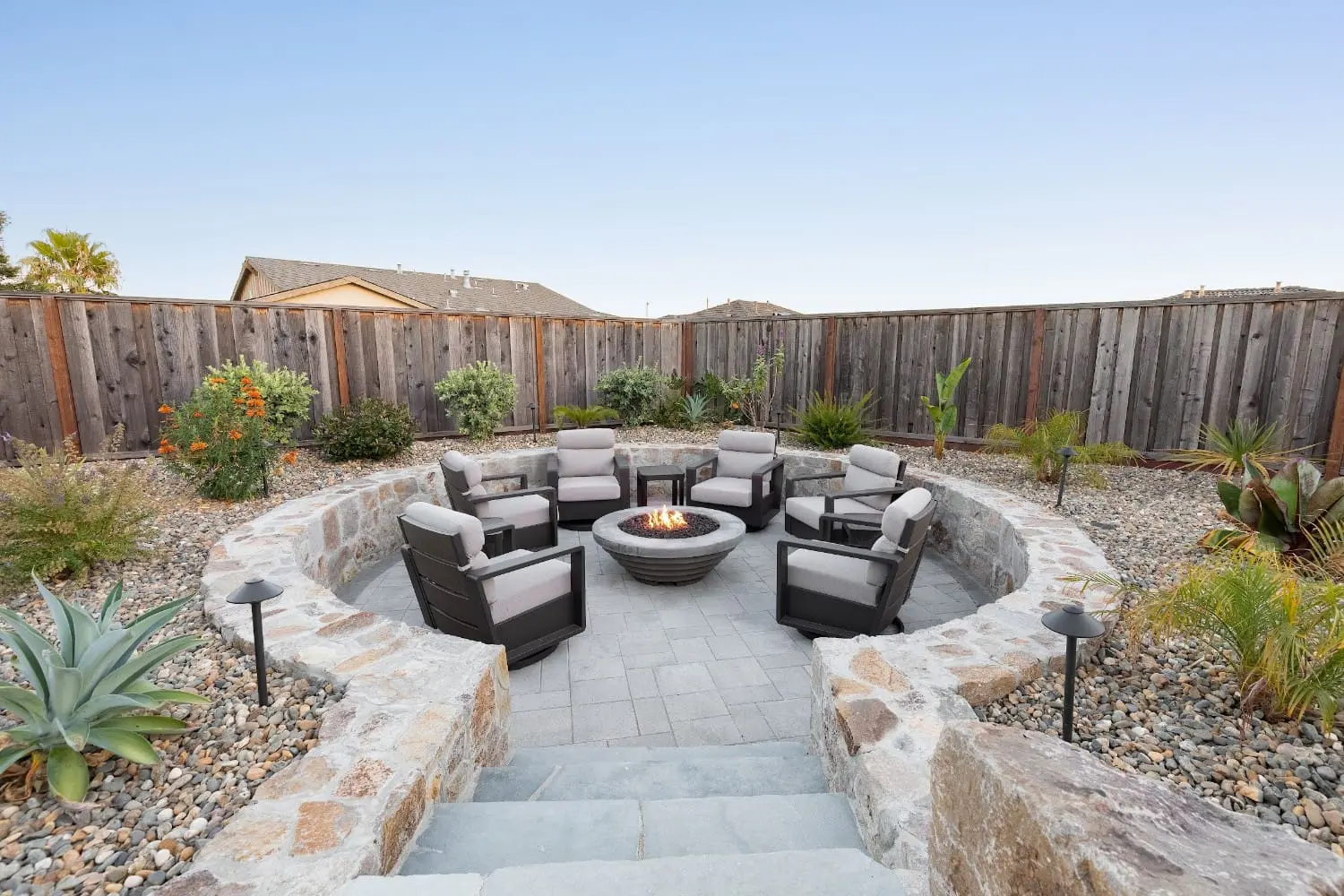 Sunken stone-walled fire pit patio with six lit gray armchairs around a lit fire bowl, surrounded by river rock beds and desert plantings, built by Keystone Yards.