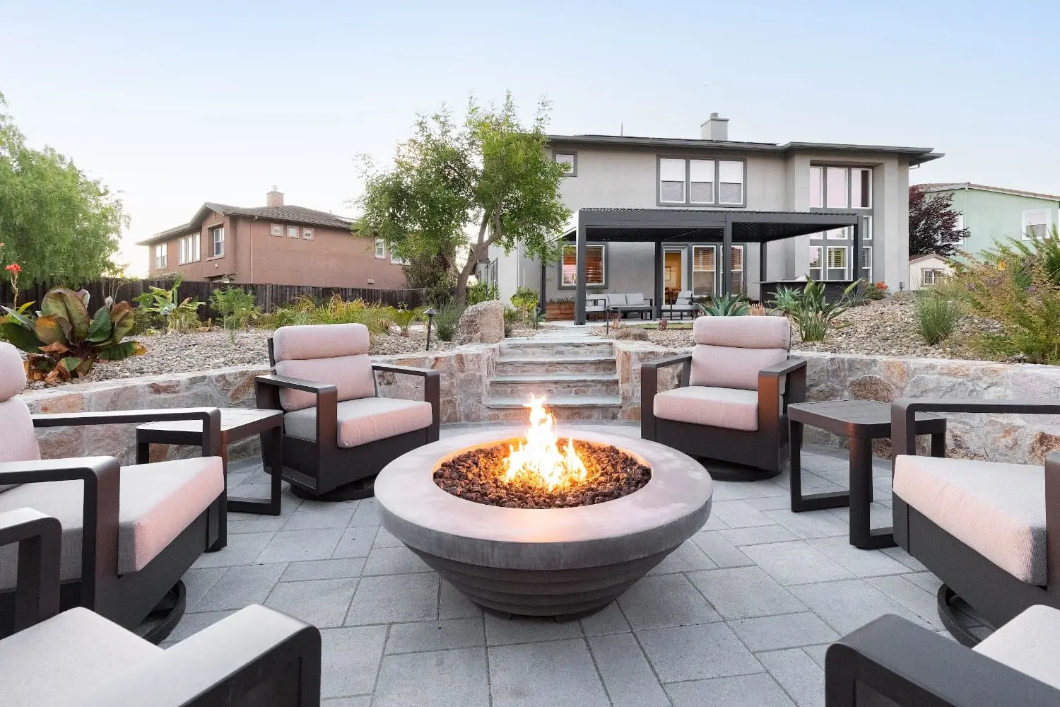 Lit fire pit surrounded by blush-cushioned lounge chairs on a paver patio, with stone-retained terraces and steps leading up to the covered back porch, by Keystone Yards.