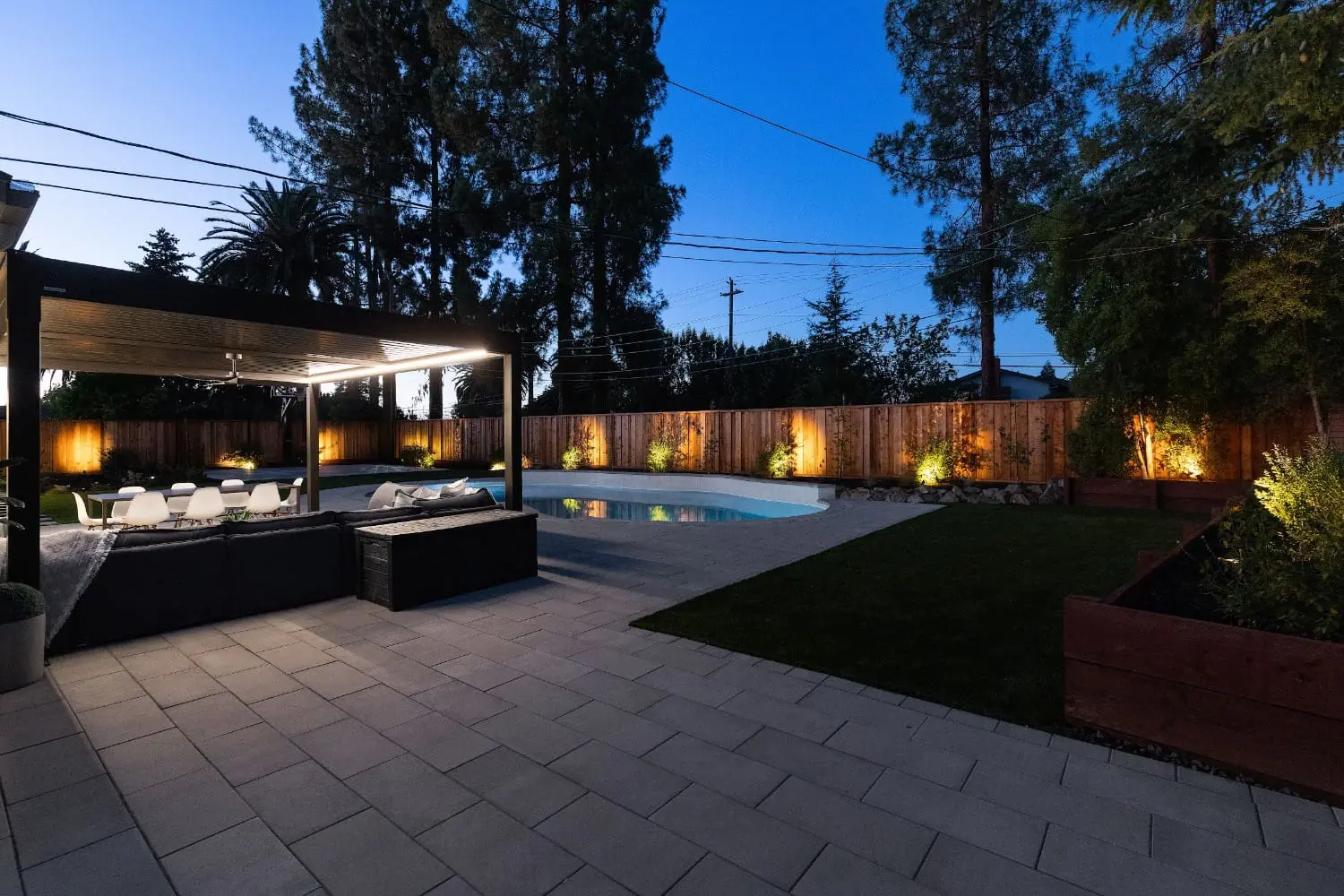 Twilight backyard by Keystone Yards with a lit pergola over a black sectional and dining set, a curved pool with uplit cedar fence, turf lawn, and raised cedar planter beds.