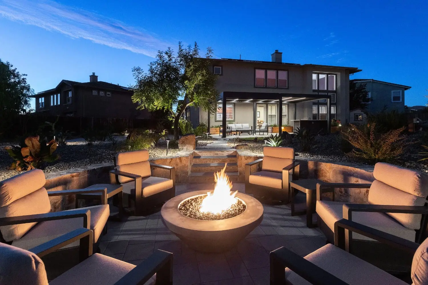 Twilight view of a sunken stone-walled fire pit lounge by Keystone Yards, with six lit lounge chairs around the flame bowl, stone steps up to a covered patio, and desert planting beds.
