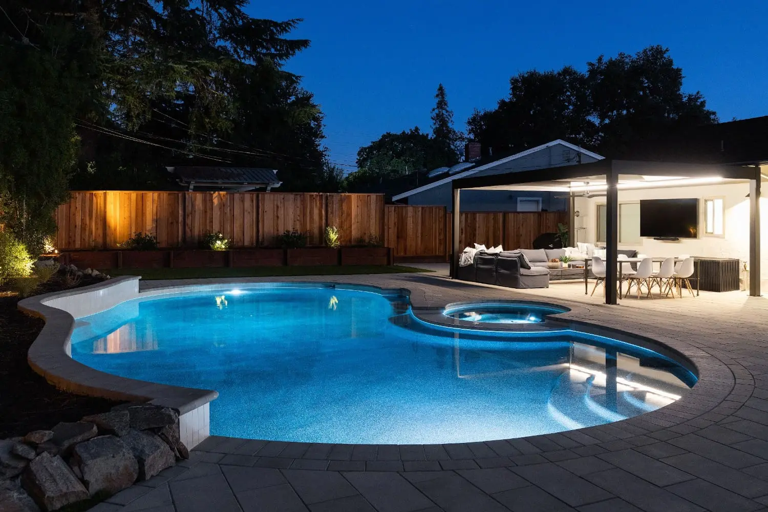 Twilight view of a freeform lit pool and spa by Keystone Yards, with a paver patio, boulder edging, cedar planter beds, and a covered lounge and dining patio.