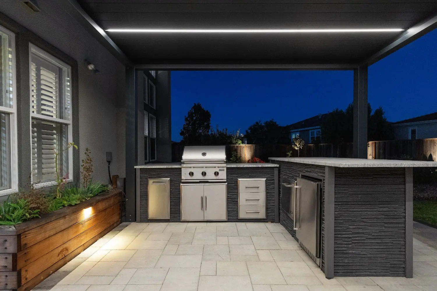 Twilight view of an L-shaped outdoor kitchen by Keystone Yards, with a stainless steel grill, storage drawers, wine fridge, and stone countertop under a lit pergola.
