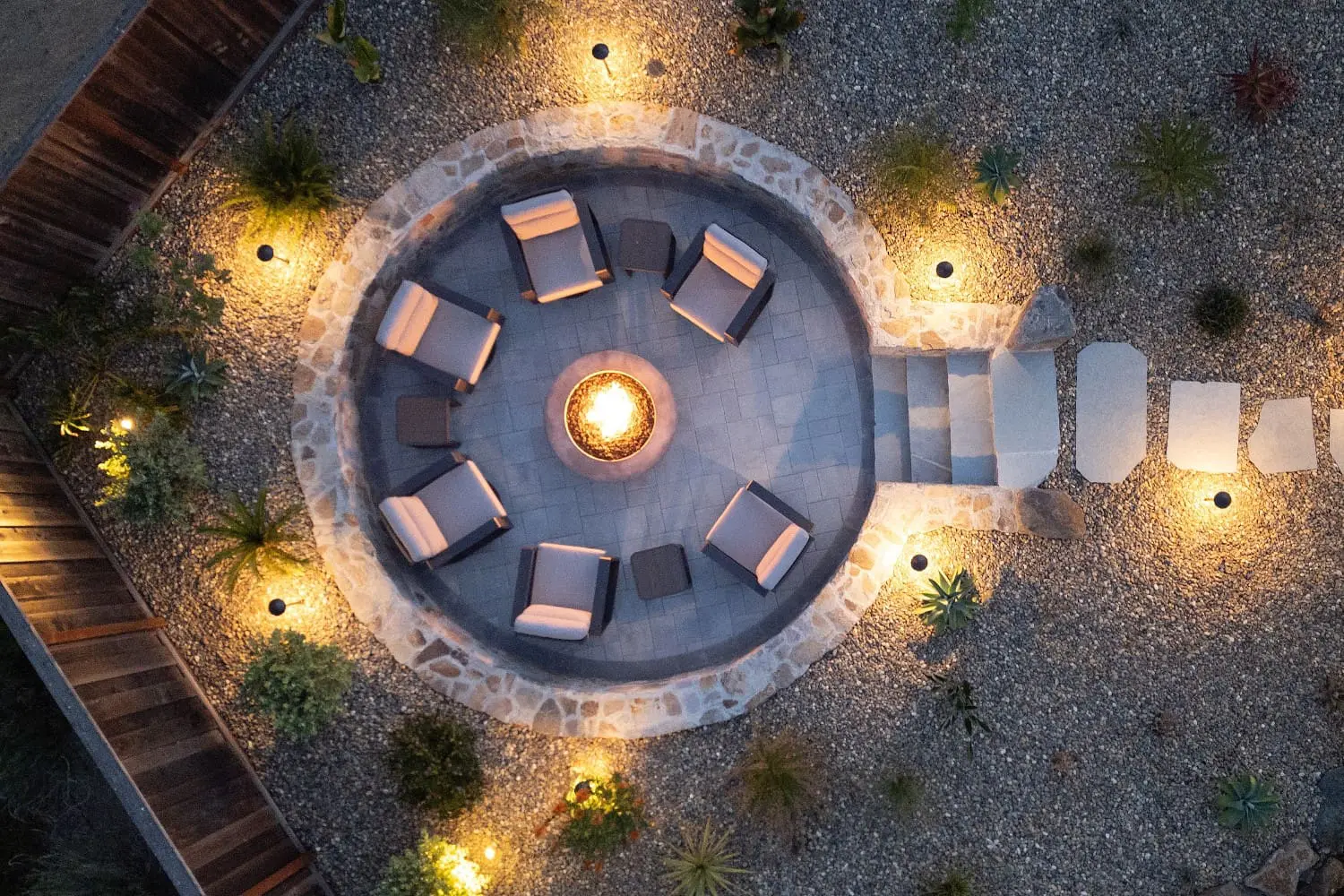 Aerial night view of a sunken stone-walled fire pit patio by Keystone Yards, with six lounge chairs lit around the flame, landscape lighting, and a stone stepping-stone path through gravel.