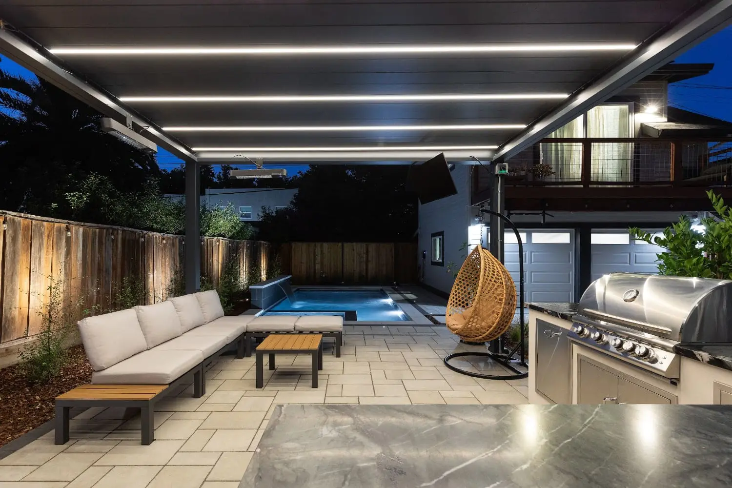 Twilight view under a lit pergola by Keystone Yards, with a white sectional sofa, TV, hanging rattan chair, stainless steel outdoor kitchen, and a lap pool with water feature.