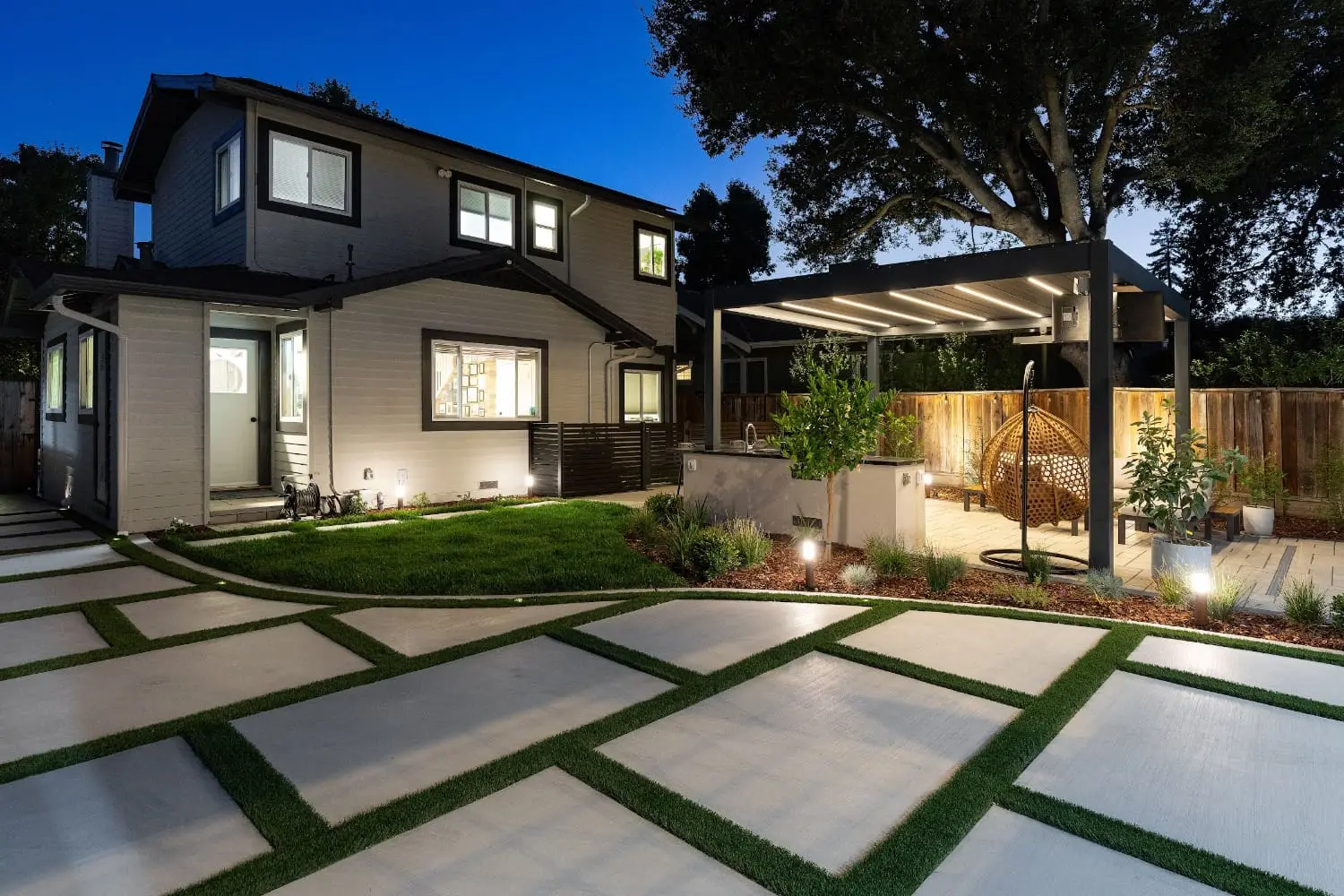 Twilight view of a backyard by Keystone Yards with a lit pergola over a hanging rattan chair and outdoor kitchen, a checkerboard concrete-and-turf pathway, and landscape lighting along the beds.