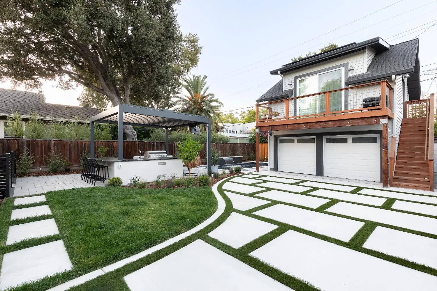 Backyard by Keystone Yards with a geometric concrete-and-turf paver driveway, a pergola-covered outdoor kitchen with barstool seating, a lawn, and a pool with a stone waterfall by the two-story garage.