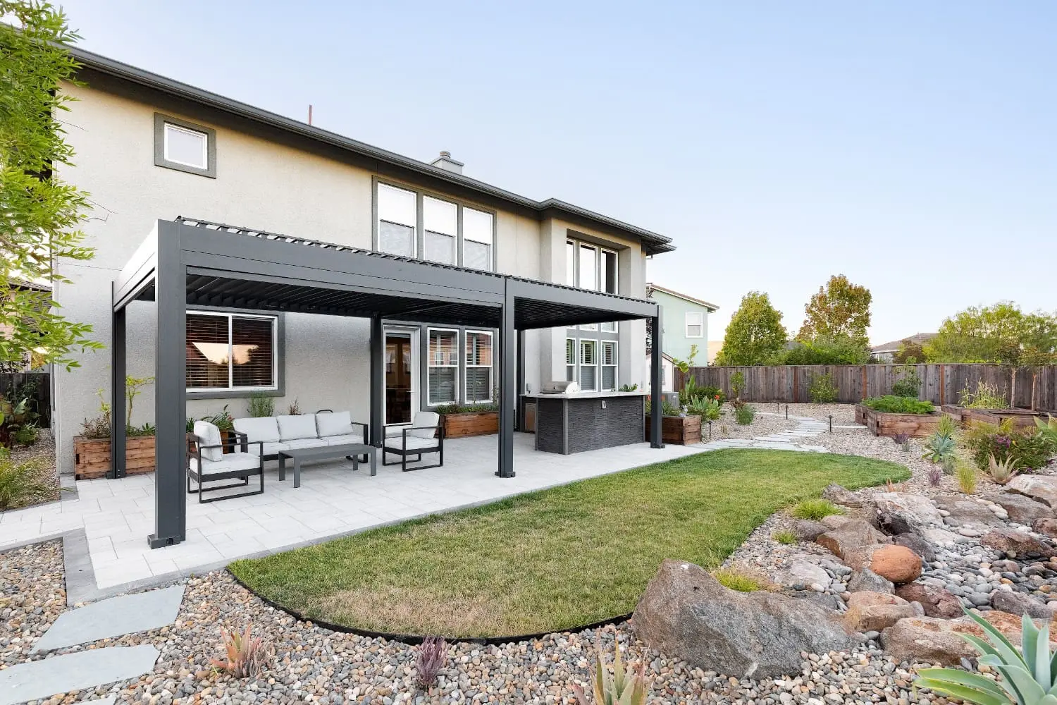 Backyard by Keystone Yards with a black louvered pergola over a lounge and outdoor kitchen patio, a curved turf lawn, and a dry riverbed rock garden with agave and succulents.