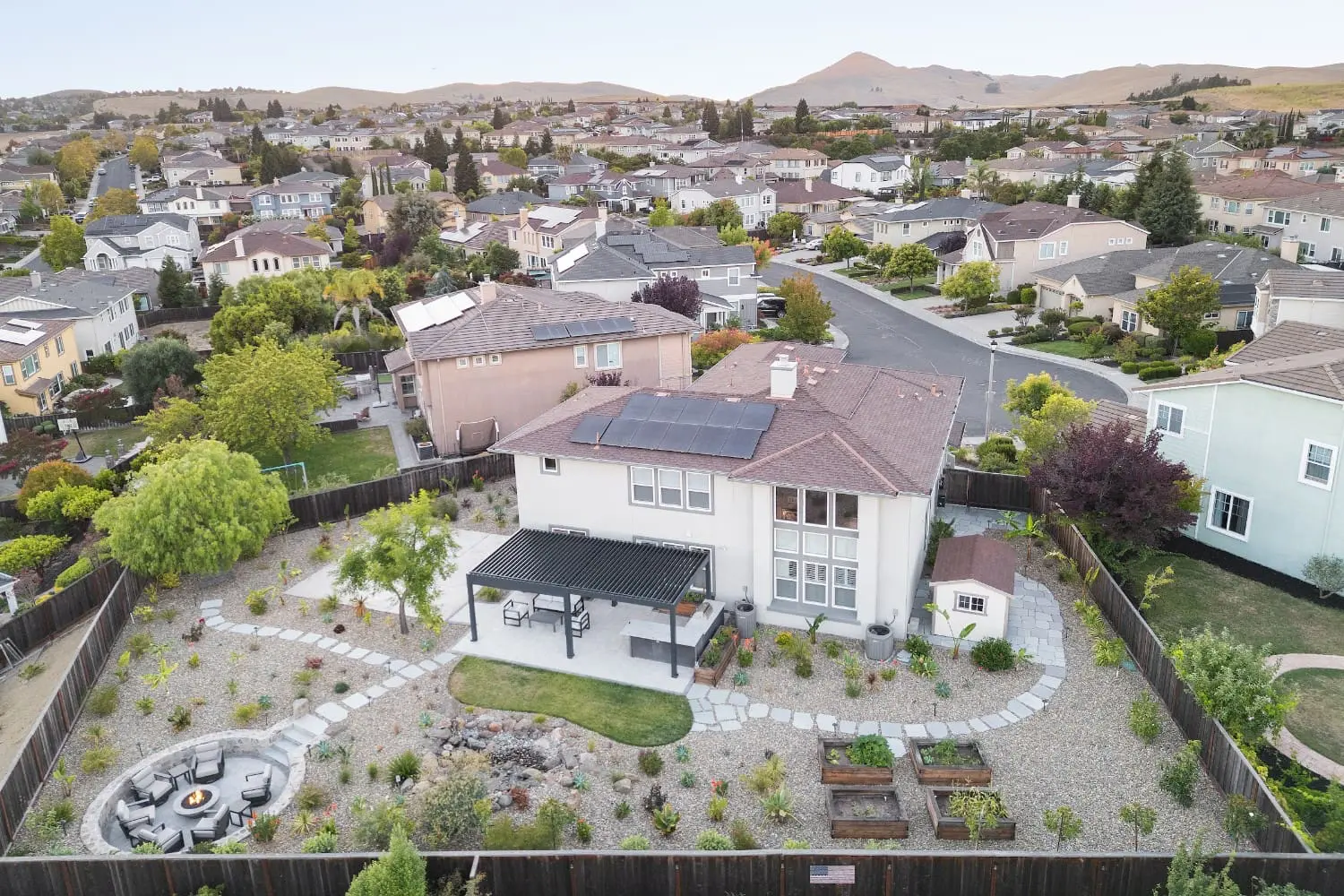 Aerial view of a suburban backyard by Keystone Yards with a solar-paneled home, pergola-covered patio, raised garden beds, a circular gravel fire pit lounge, and desert-style planting with hills in the background.