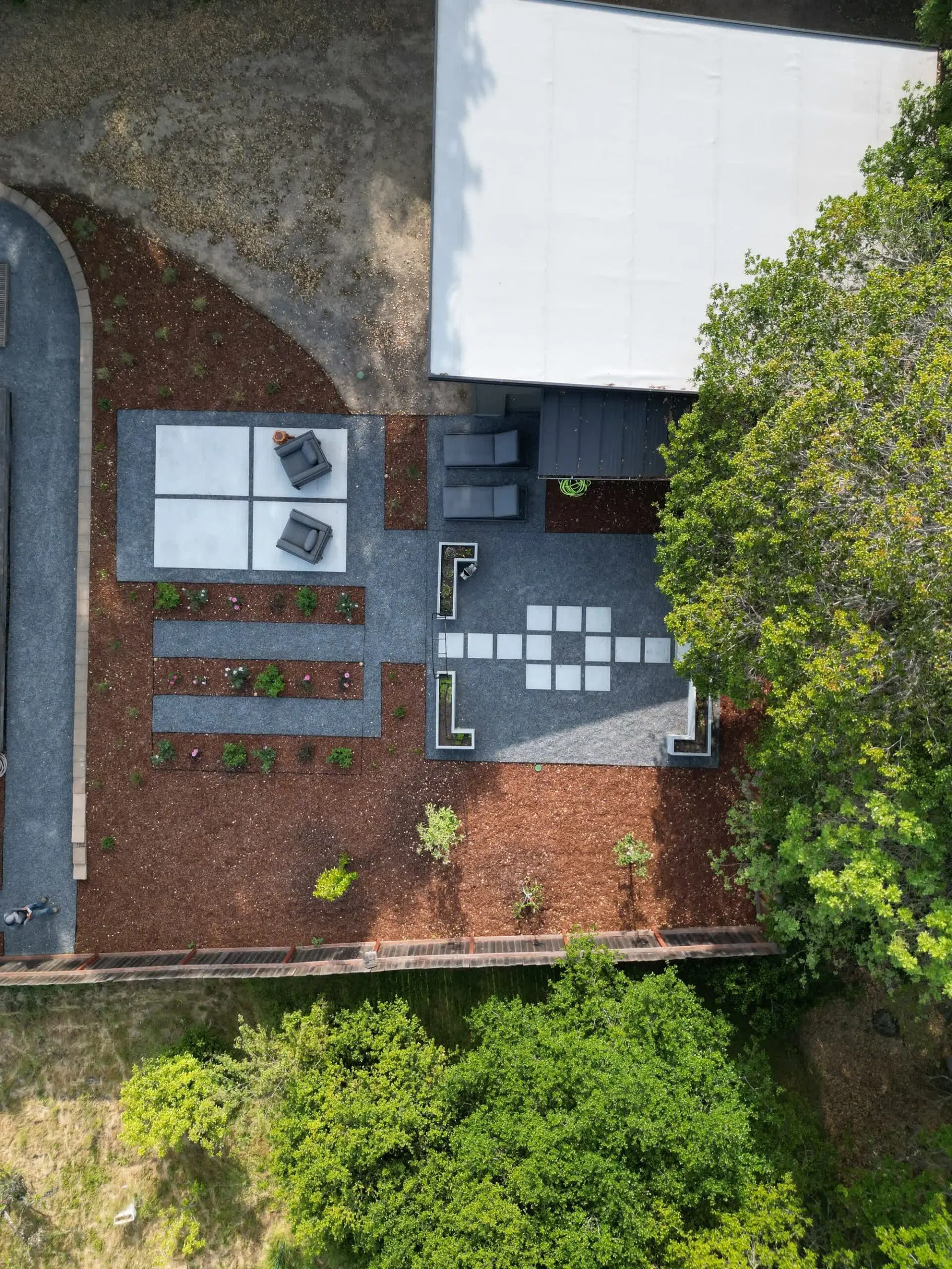 Aerial view of a backyard by Keystone Yards with gravel patios, a checkerboard paver path, raised garden beds, rose planting rows, and mulched borders beside a barn-style building.