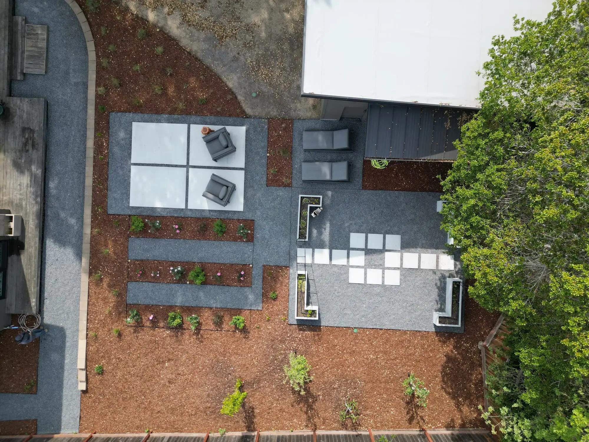 Aerial view of a modern backyard by Keystone Yards with concrete-and-gravel patios, lounge chairs, rows of rose planting beds, and a raised garden arbor beside a mature oak tree.