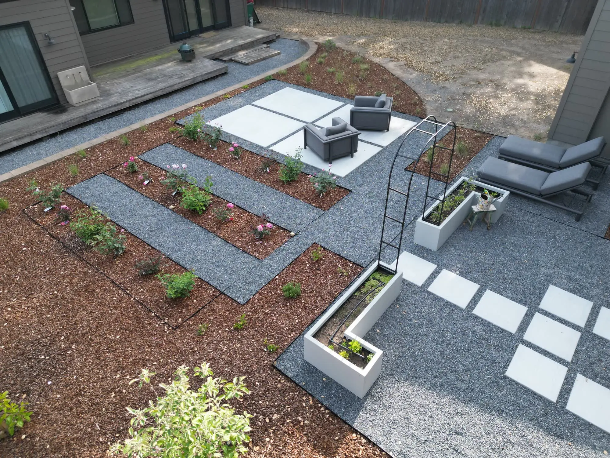 Aerial view of a modern backyard by Keystone Yards with geometric concrete-and-gravel patios, white raised planter beds, a metal garden arbor, and rows of rose planting beds in mulch.