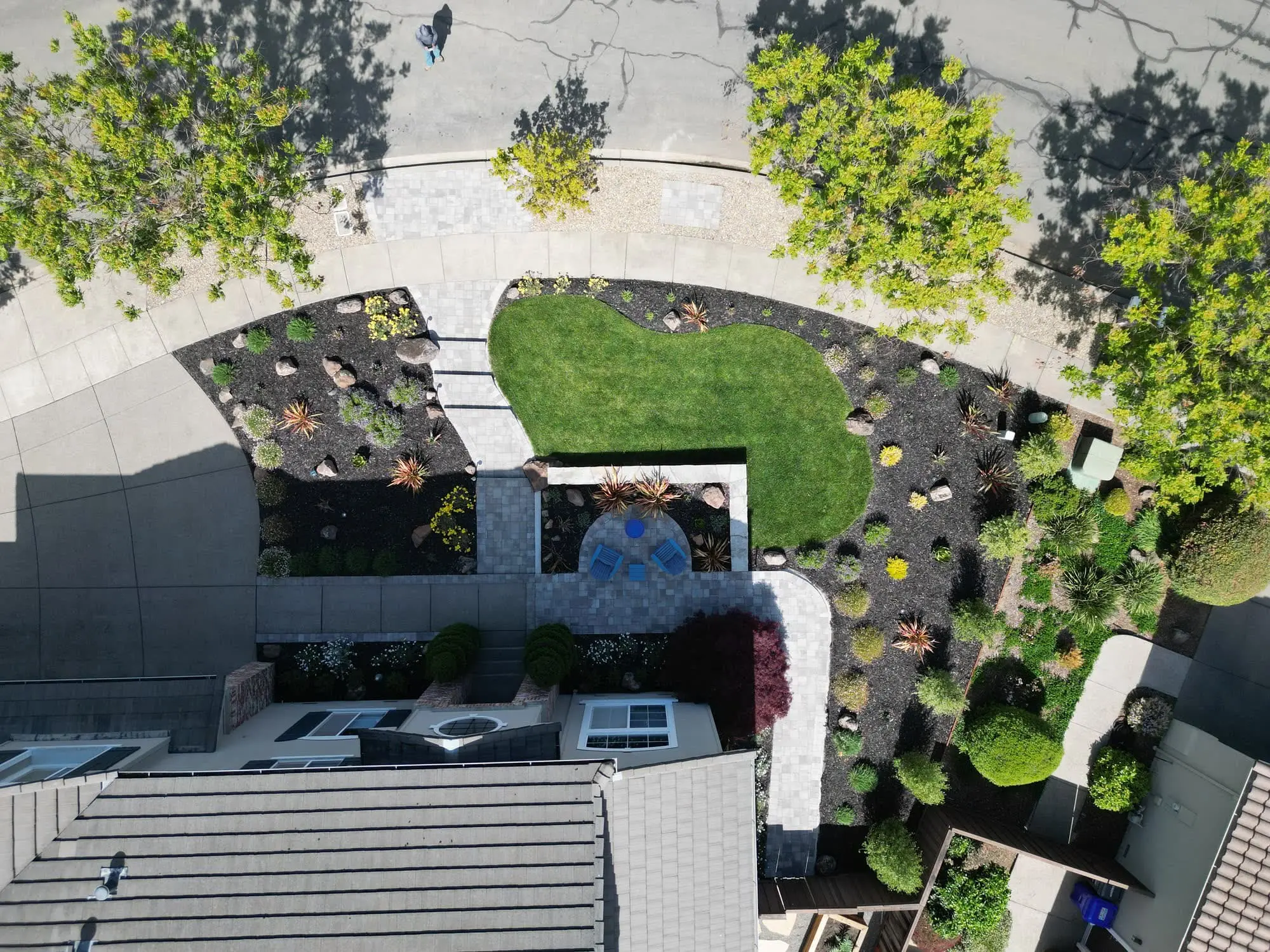 Aerial view of a curved backyard by Keystone Yards with a turf lawn, boulder-and-succulent planting beds, a round patio with blue chairs, and a paver walkway to the house.
