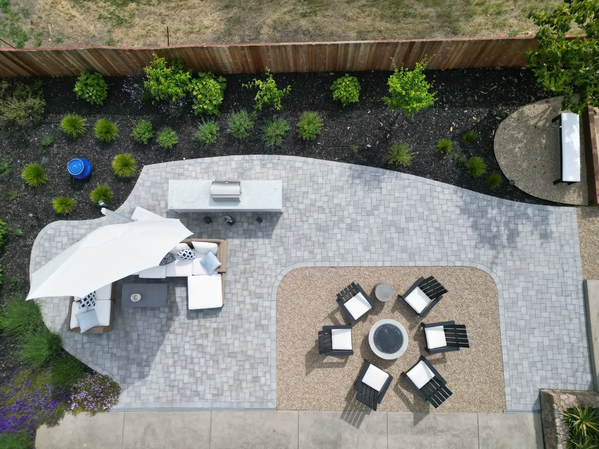 Aerial view of a curved paver patio by Keystone Yards with a built-in grill counter, umbrella-shaded sectional sofa, and a gravel fire pit lounge with six black chairs, bordered by planting beds.