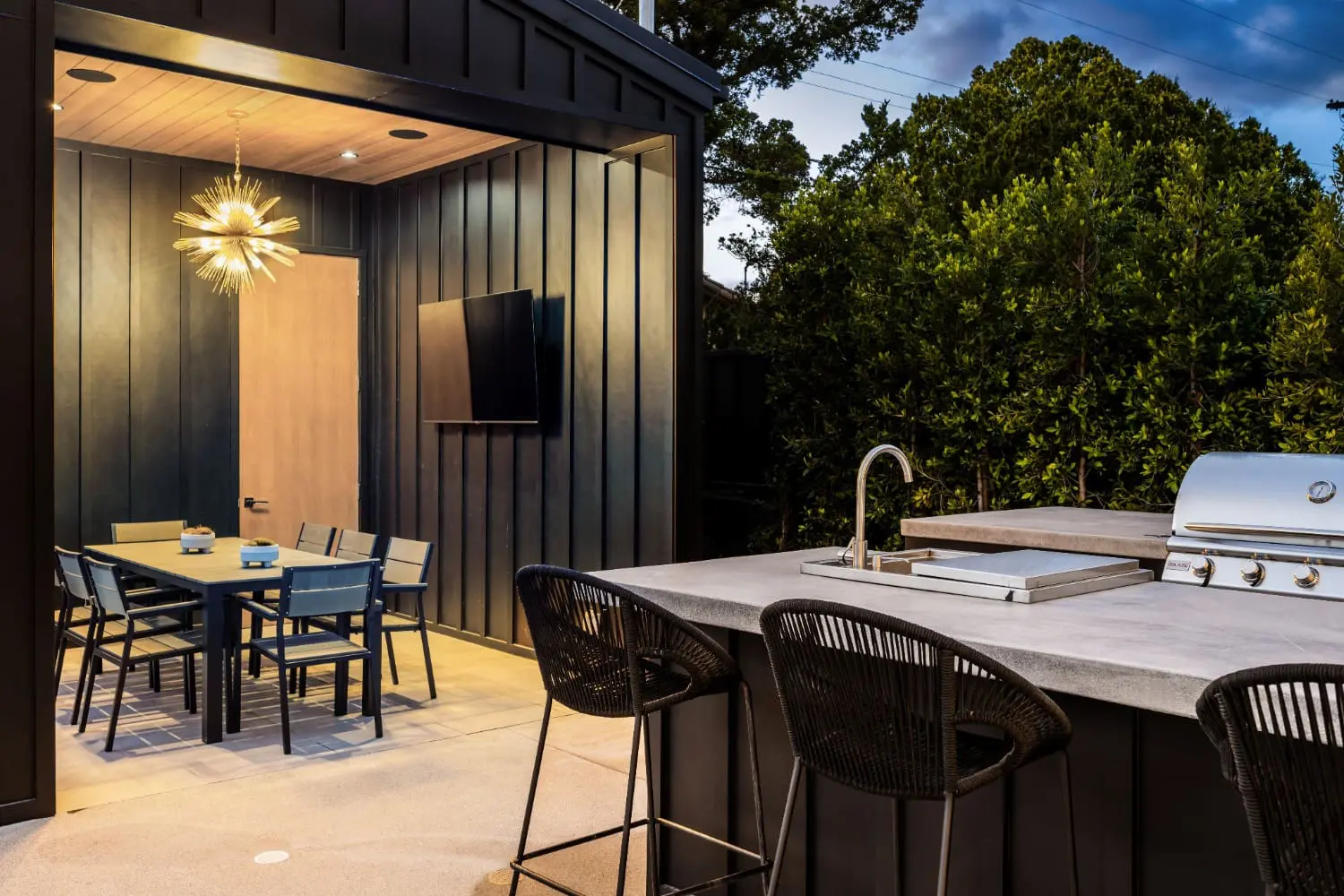 Twilight view of an outdoor kitchen and enclosed dining pavilion by Keystone Yards, with a concrete bar counter, built-in grill, woven barstools, and a black-paneled dining room lit by a starburst chandelier.