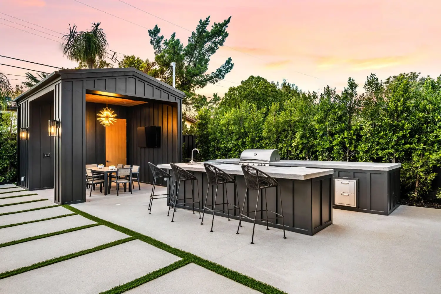 Sunset view of a black board-and-batten outdoor kitchen pavilion by Keystone Yards, with a concrete bar counter, built-in grill, woven barstools, dining area, and a paver-and-turf patio.