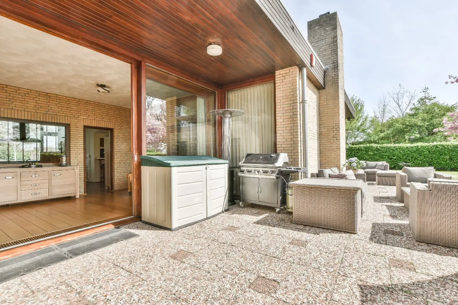 Covered patio with a wood-plank ceiling, gas grill, storage cabinet, and wicker lounge seating on a stone paver patio, an example of an outdoor kitchen in Napa by Keystone Yards.