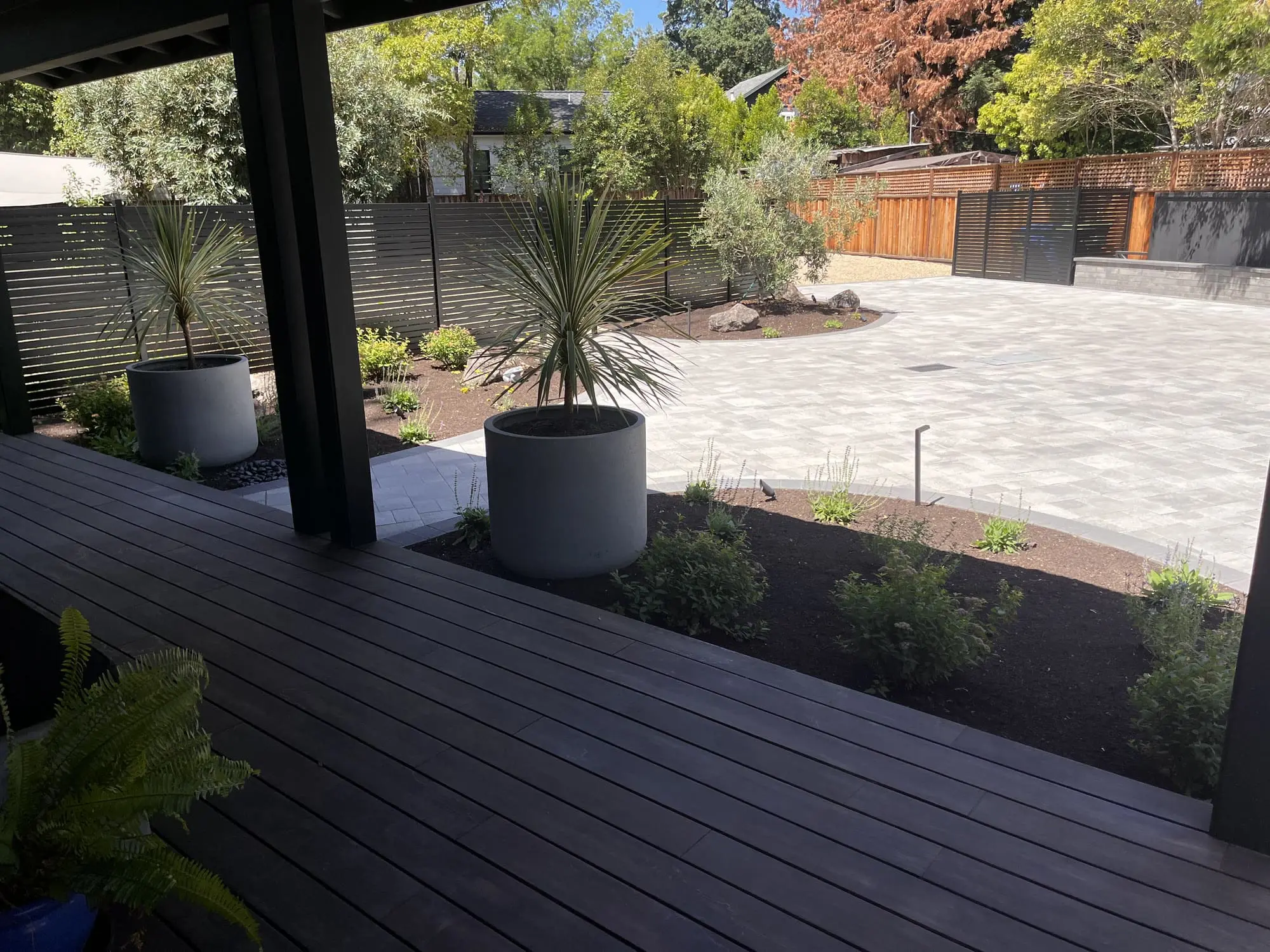 View from a dark wood deck with potted dracaena plants over a paver patio, mulched planting beds, and a modern slatted privacy fence, landscaped by Keystone Yards.