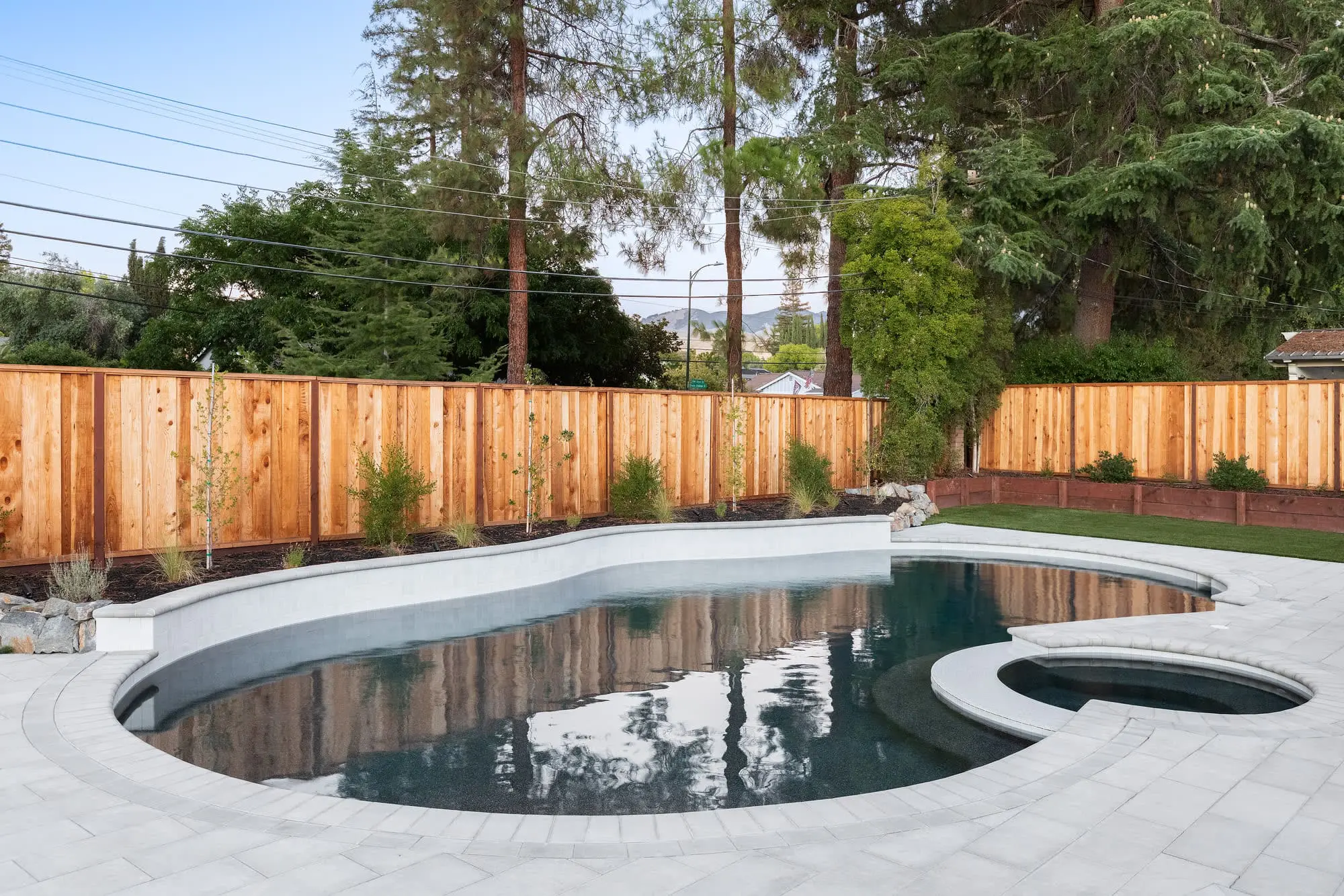 Freeform kidney-shaped pool with an attached round spa and dark plaster finish, bordered by a paver patio and cedar privacy fence with new planting beds, built by Keystone Yards.