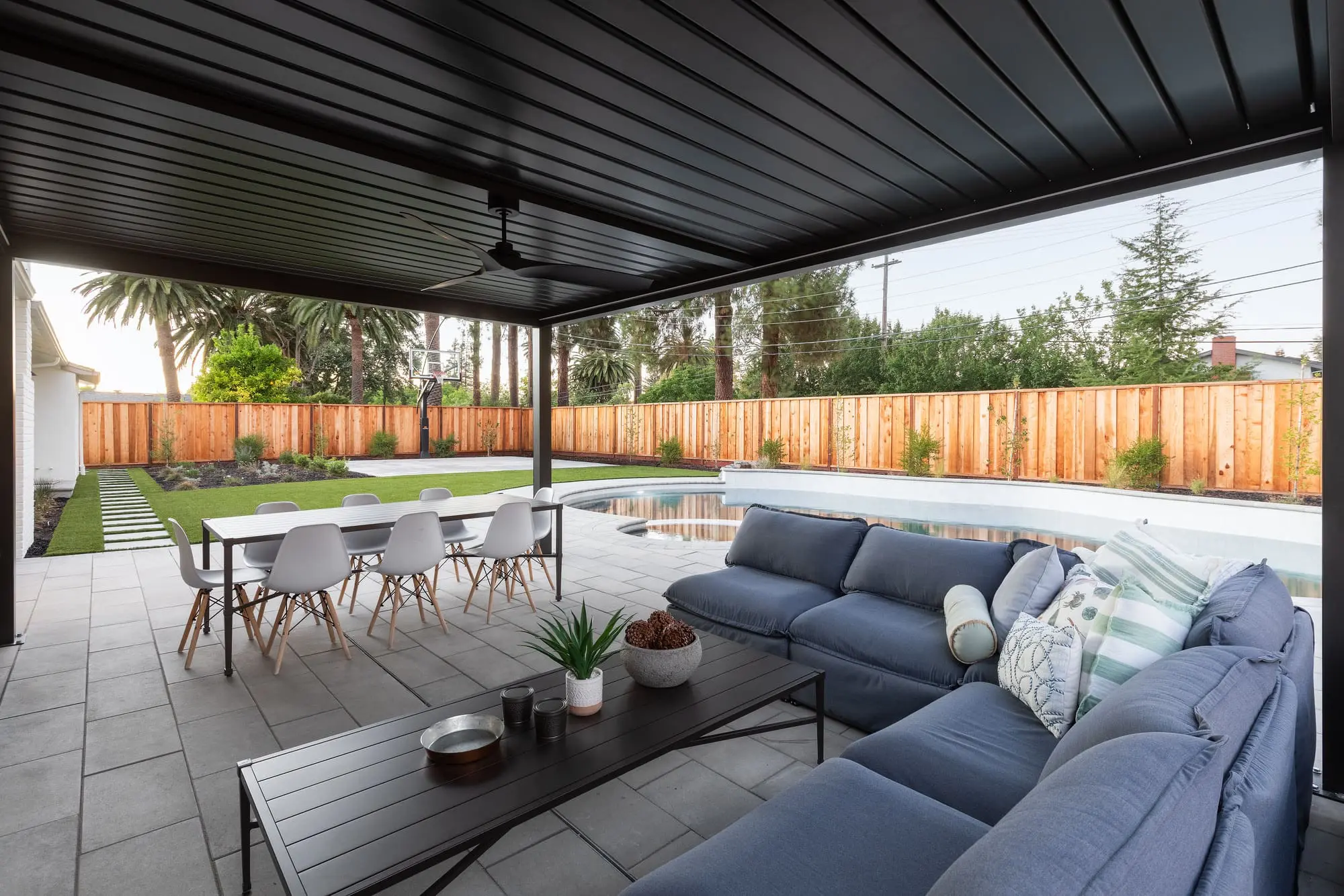 Covered patio by Keystone Yards with a black louvered pergola over a blue sectional sofa and dining table, overlooking a curved pool, turf lawn, basketball hoop, and cedar privacy fence.
