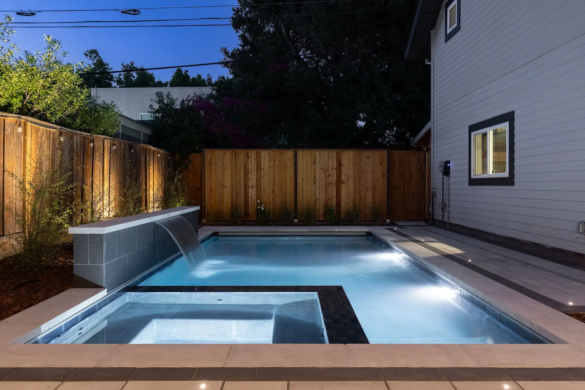 Twilight view of a lit pool and spa with a raised stone waterfall wall, uplit wood privacy fence, and paver patio, built by Keystone Yards.