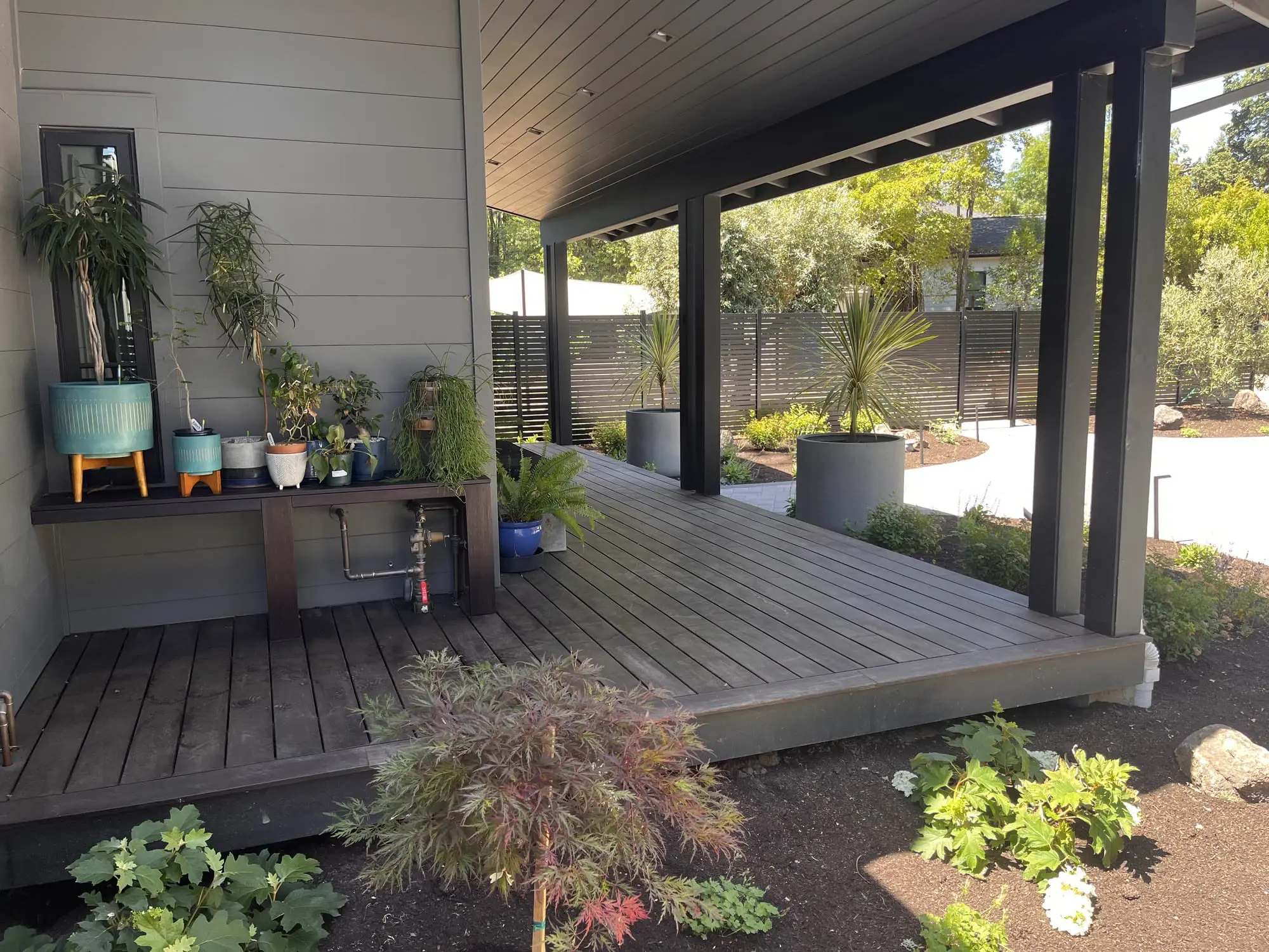 Covered dark wood deck with a potting bench of houseplants and a Japanese maple, opening onto a slatted privacy fence and mulched planting beds, landscaped by Keystone Yards.