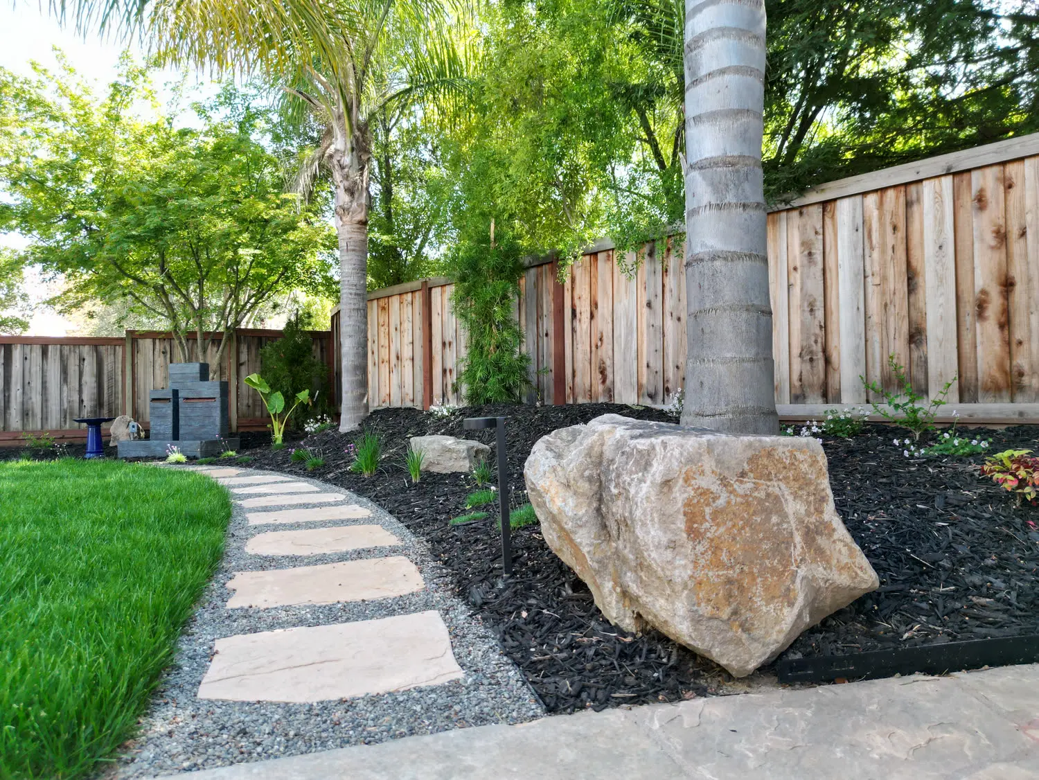 tone paver walkway curved through a lush green lawn, bordered by dark mulch, a large natural accent boulder, and wooden fencing.