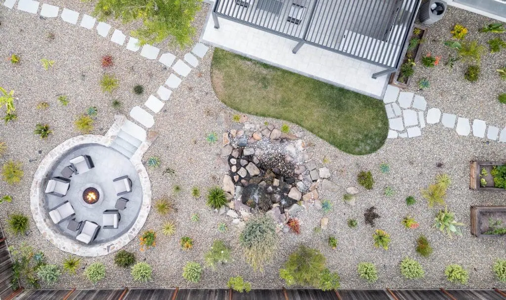 Bird's-eye view of a desert-style backyard by Keystone Yards, with a circular stone fire pit lounge, a rock water feature, a curved patch of lawn, and gravel grounds dotted with