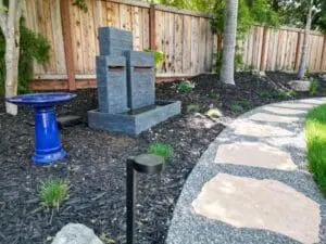 Tiered slate water fountain with a blue ceramic birdbath beside a flagstone stepping-stone path through dark mulch landscaping, designed by Keystone Yards.