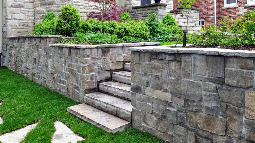 Natural stone retaining wall with built-in steps supporting a raised garden bed in a luxury landscape.