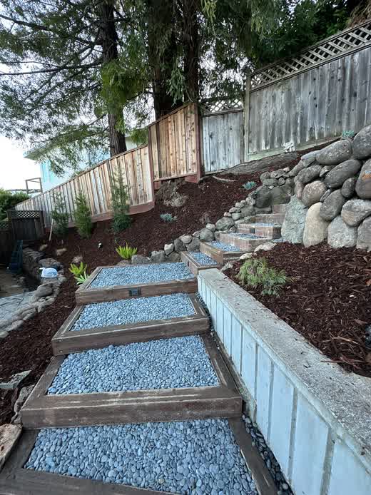 Terraced hillside steps with wood-framed blue river rock treads climbing past boulder retaining walls and mulched planting beds, built by Keystone Yards.