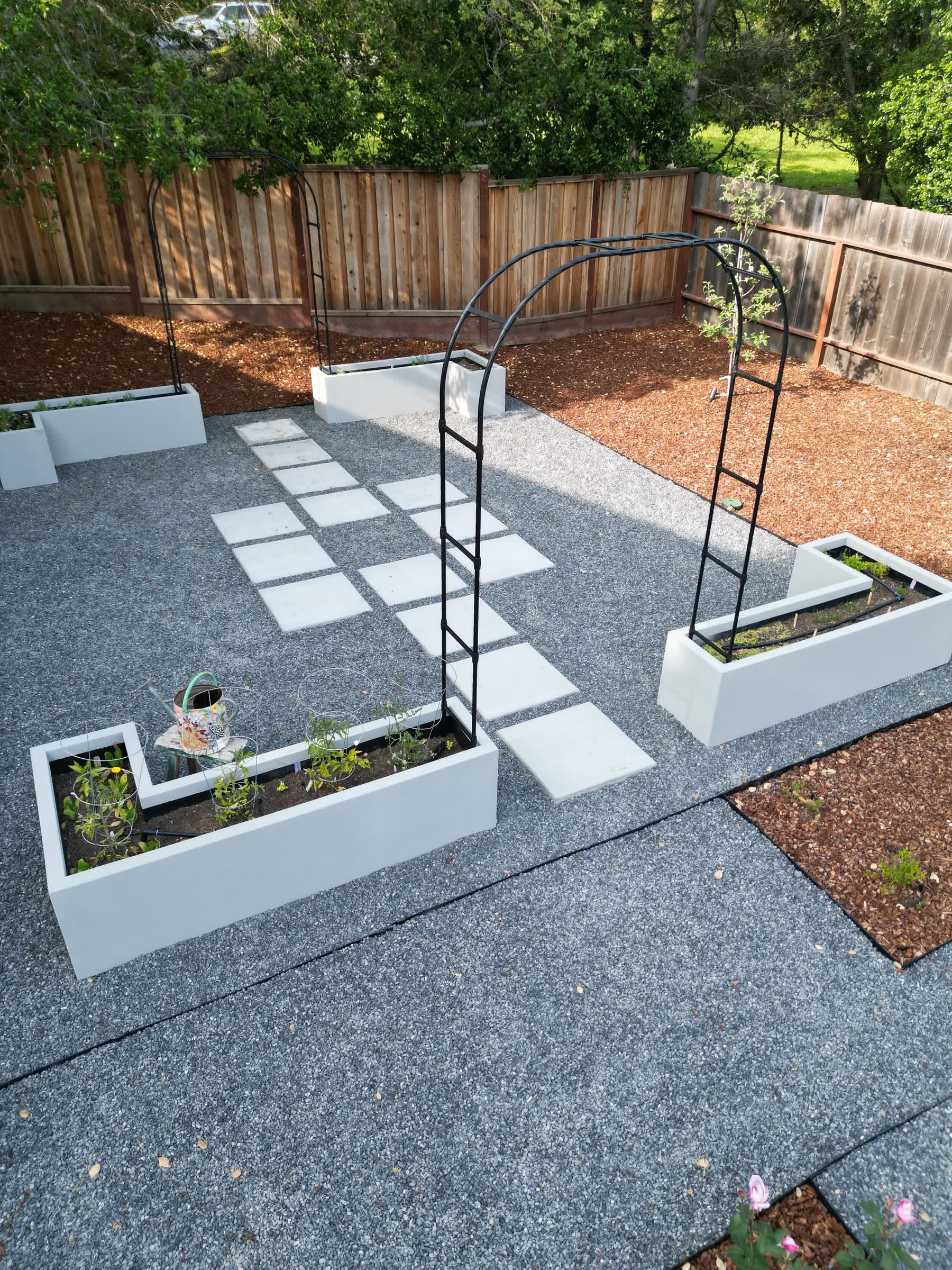 White L-shaped raised garden beds with tomato cages and metal arbors, set on a gravel patio with a checkerboard paver path, bordered by mulch and a wood fence, built by Keystone Yards.
