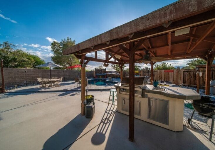 Covered wood pergola with outdoor kitchen, concrete patio, pool, and dining area in a complete backyard entertainment space