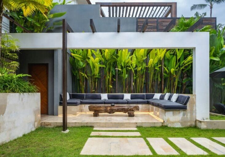 Contemporary outdoor lounge with built-in sectional seating, rustic wood coffee table, tropical plant wall, and stone pathway