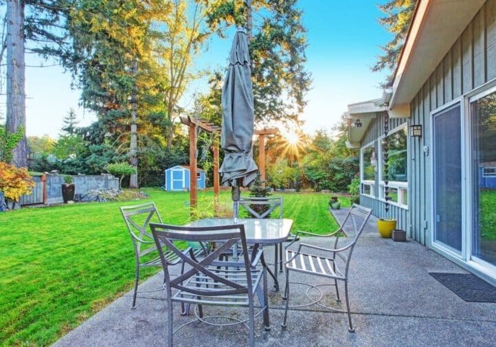 Backyard patio with metal dining set, lush striped lawn, wood pergola, garden shed, and golden sunset through tall trees
