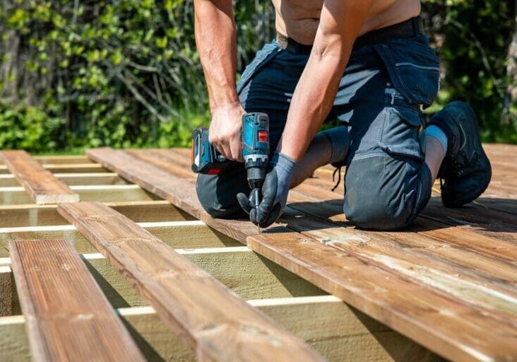 Professional carpenter installing wood deck boards with a power drill during outdoor deck construction