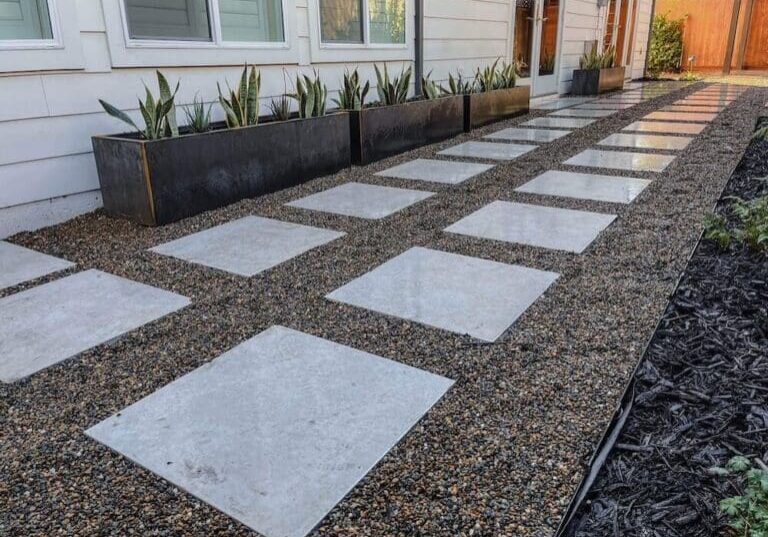 Modern front yard with large concrete pavers, gravel ground cover, and black metal planters with succulents