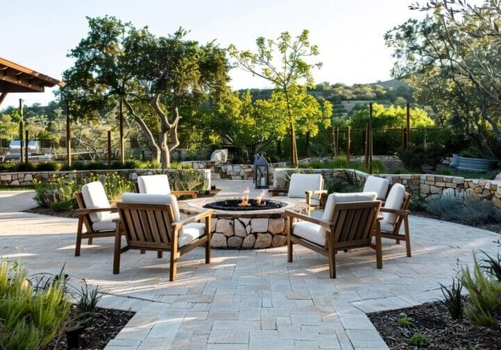 Round stone fire pit with teak lounge chairs, travertine patio, stone retaining walls, and mature oak tree backdrop