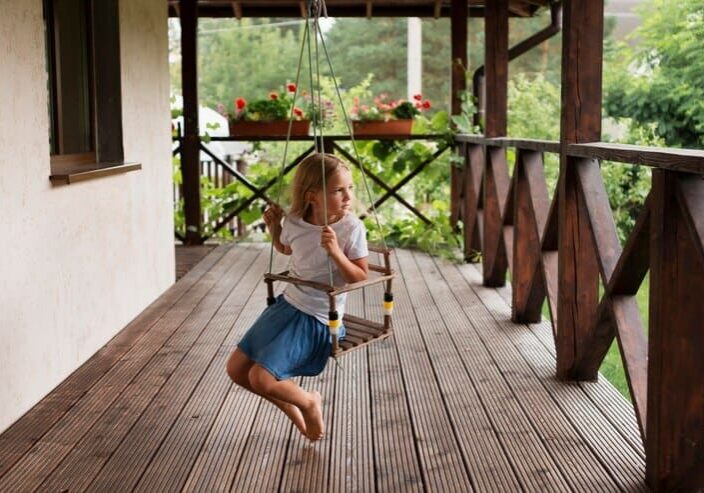 Child playing on a wooden swing on a rustic wood deck porch surrounded by lush garden greenery