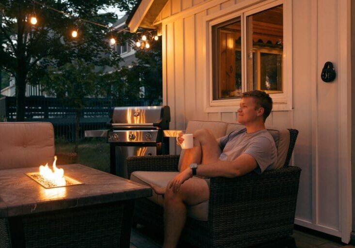 Man relaxing on a wicker patio chair beside a fire pit table and gas grill under evening string lights