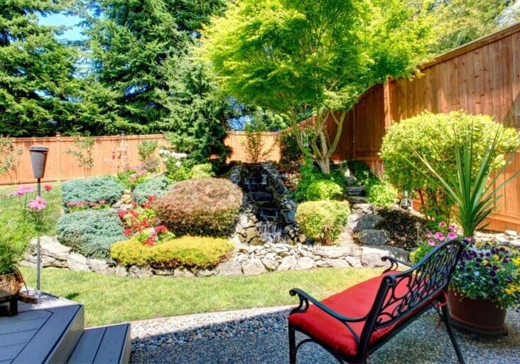 Backyard garden with stone waterfall feature, colorful shrubs, and iron bench