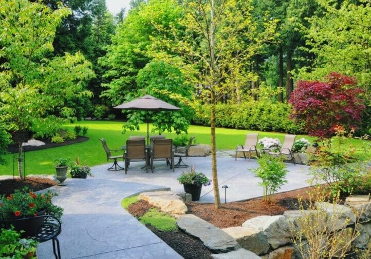 Lush backyard patio with dining set, umbrella, stone edging, and mature tree landscape