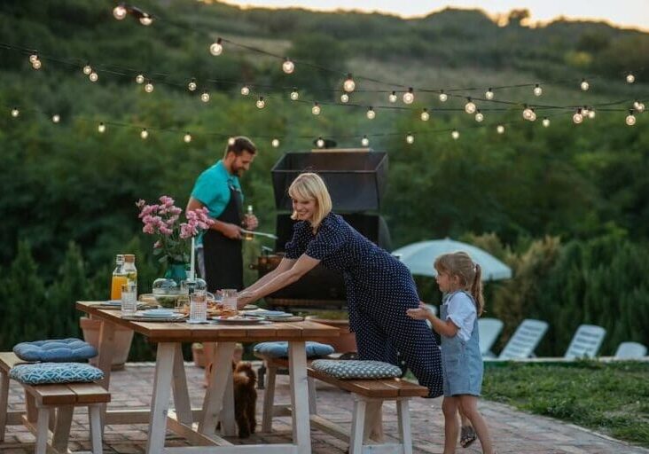 Family enjoying outdoor dining with string lights and backyard grill at dusk