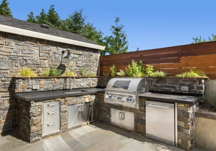 Built-in outdoor kitchen with stone masonry, stainless steel grill, black countertops, and herb garden planters