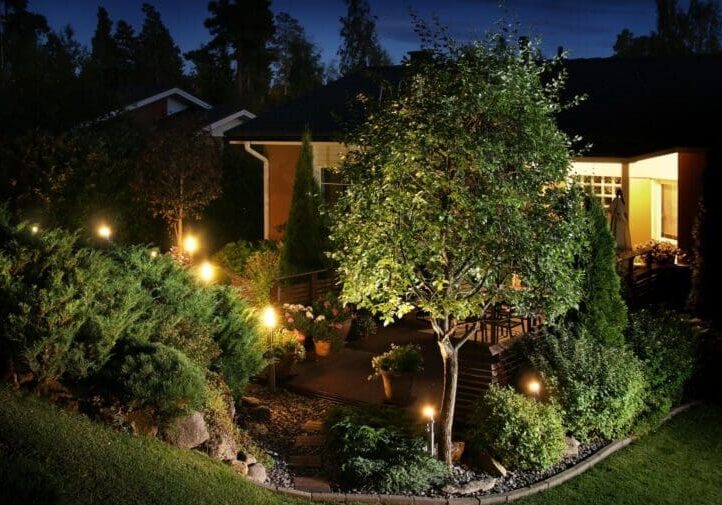 Residential backyard with landscape lighting, mature trees, and deck at night
