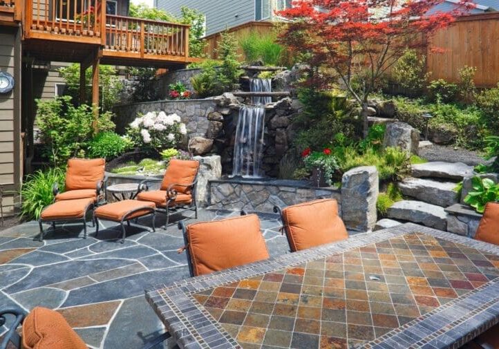 Stone patio outdoor living space with wrought iron seating, mosaic tile table, and cascading rock waterfall surrounded by lush gardens