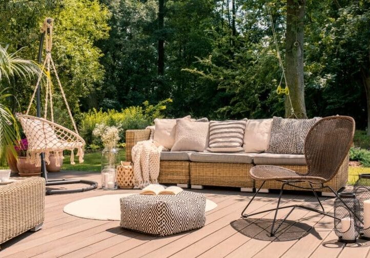 Bohemian wood deck with wicker sofa, macrame hanging chair, patterned pouf, and lanterns surrounded by lush garden trees