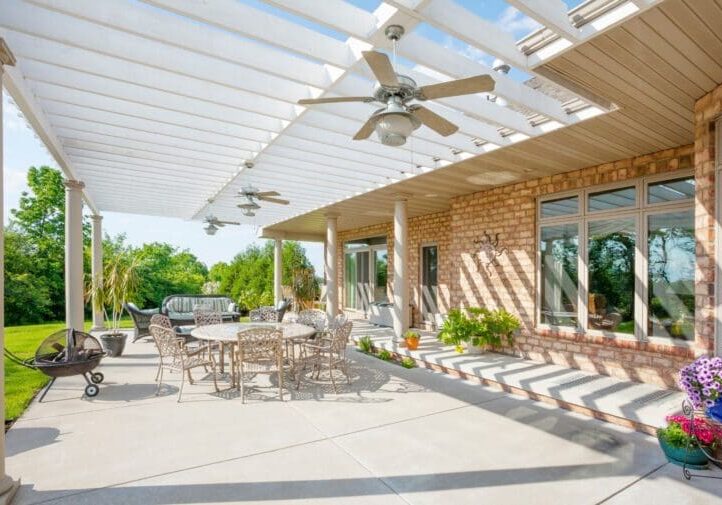 Spacious covered patio with white pergola, ceiling fans, wrought iron dining set, fire pit, and lush green backyard views